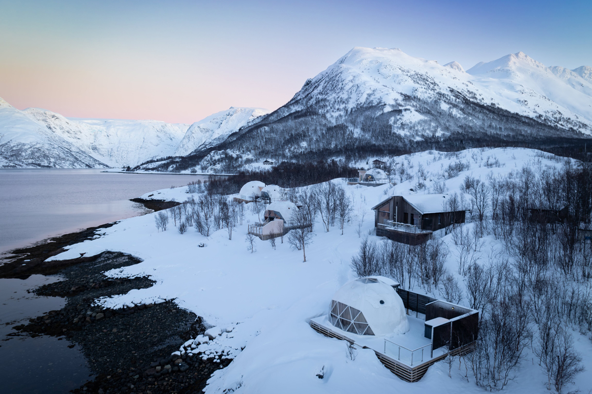 Isbreen's glass igloos and cabins along the fjord shore at dusk, surrounded by snowy peaks.