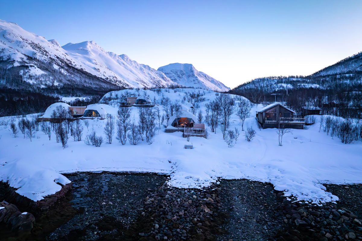 Isbreen's igloos and cabins set among snowy birch trees with mountains rising behind.