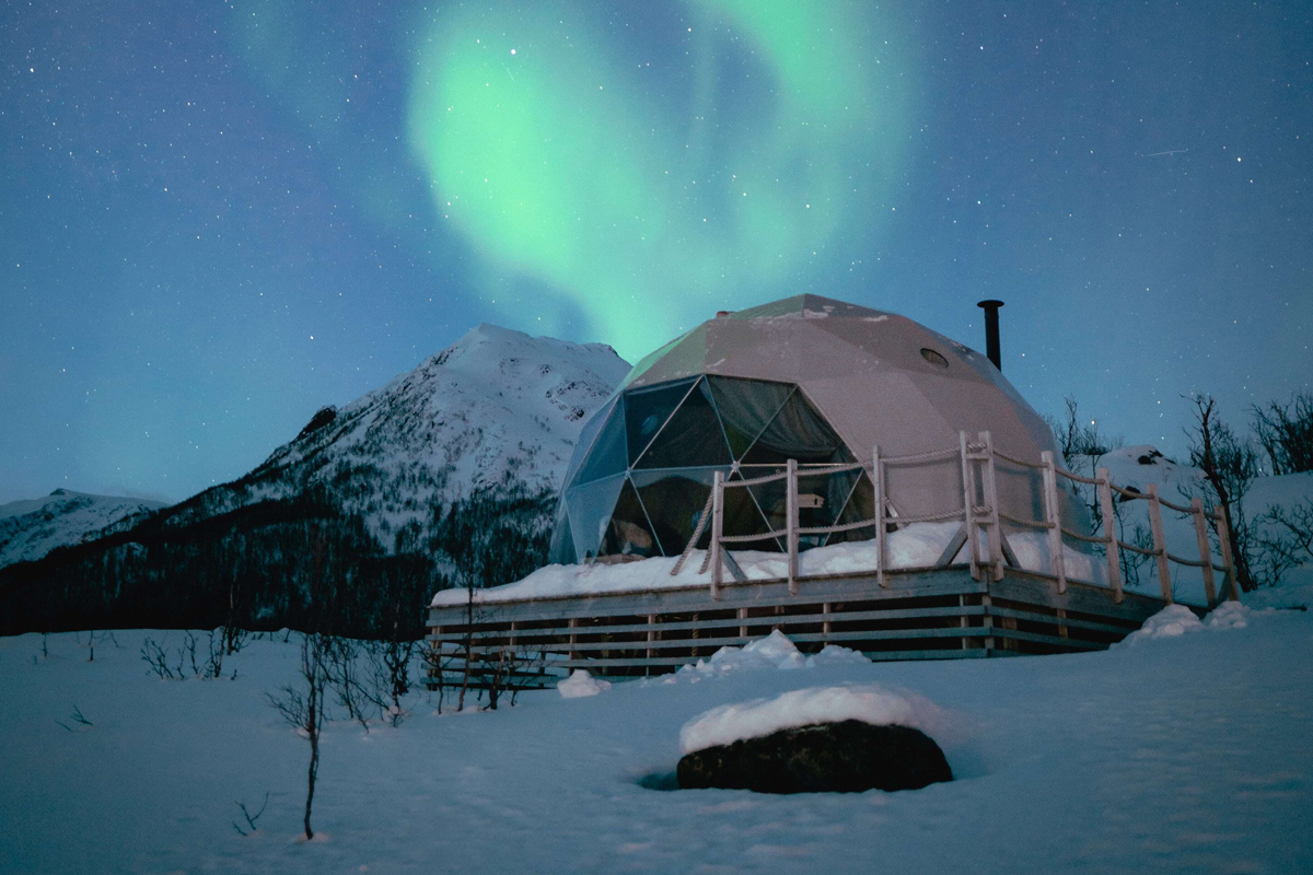 A glass igloo glows beneath the northern lights with snow-capped peaks behind at Isbreen.