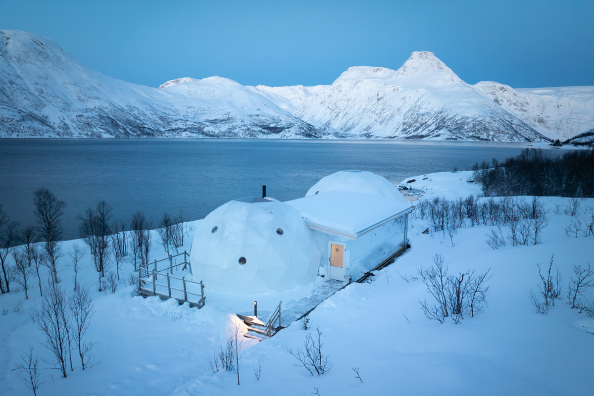 Twin igloo domes at Isbreen glowing at dusk beside a still fjord and snow-capped mountains.