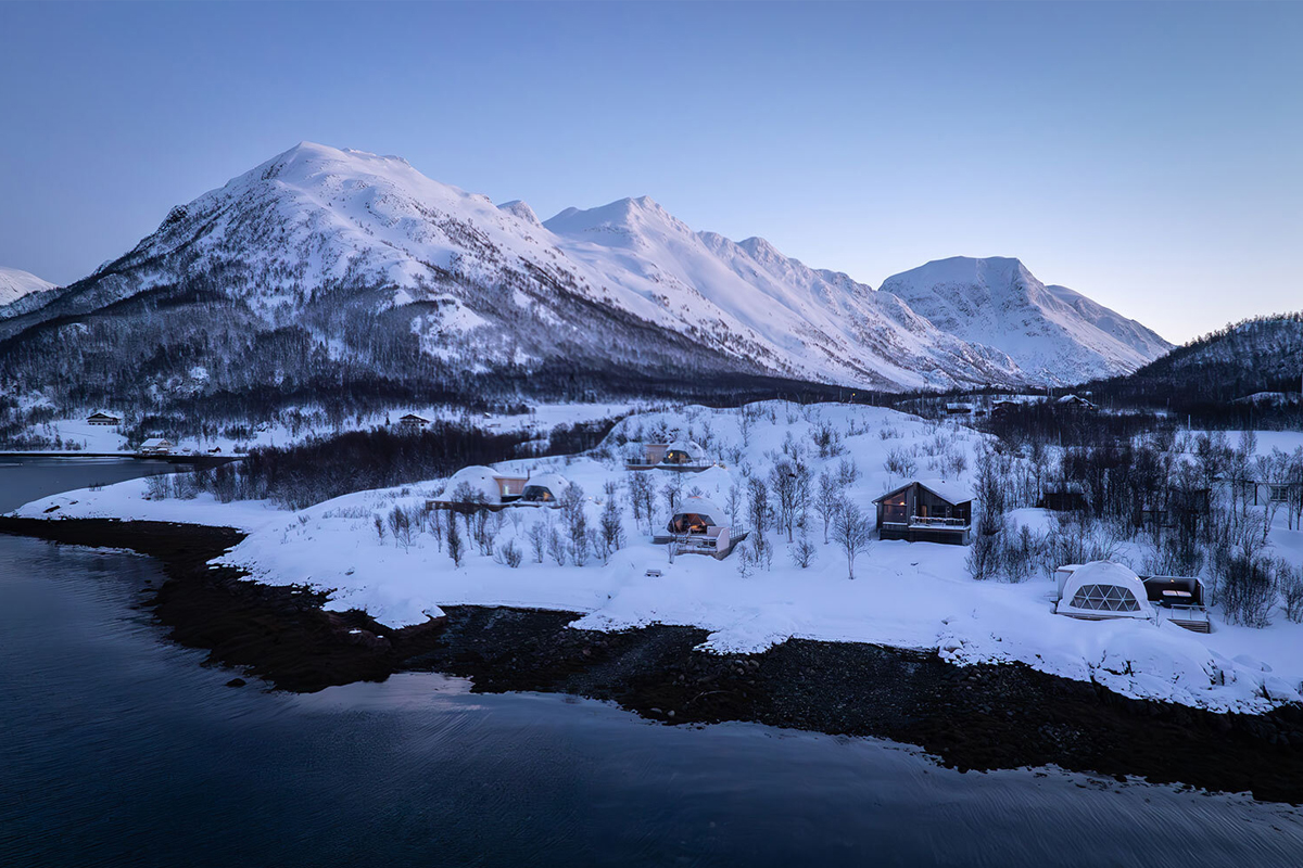 Aerial dusk view of Isbreen glass igloos and cabins along the fjord shore, framed by mountains.