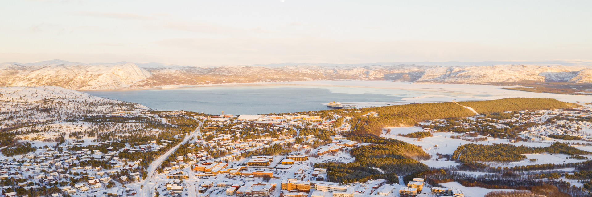 Aerial view of Alta, Norway, with snow-covered mountains framing the fjord and town below.
