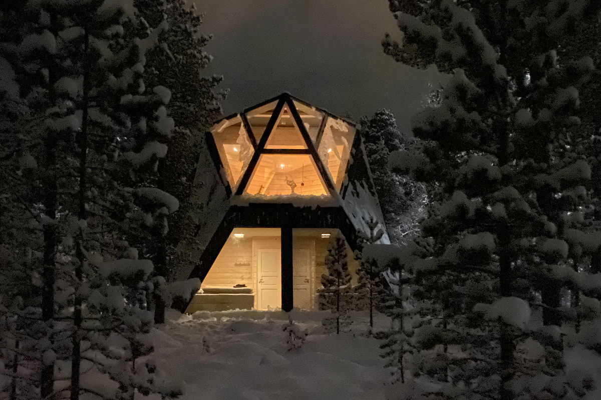 Glass cabin at Trasti & Trine glowing warmly at night, framed by snow-laden trees.
