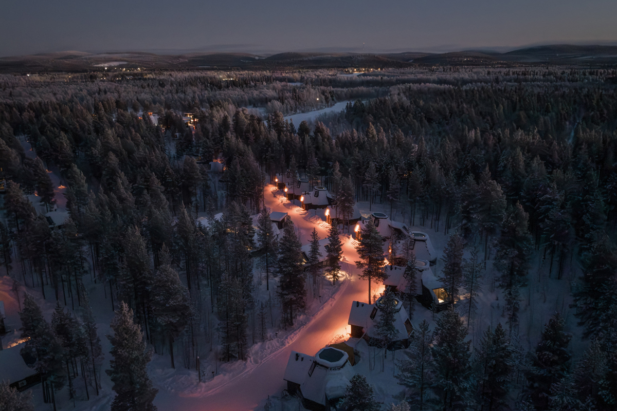 Aerial night view of Aurora Village with illuminated paths and snow-covered cabins set among frost-covered trees, Ivalo