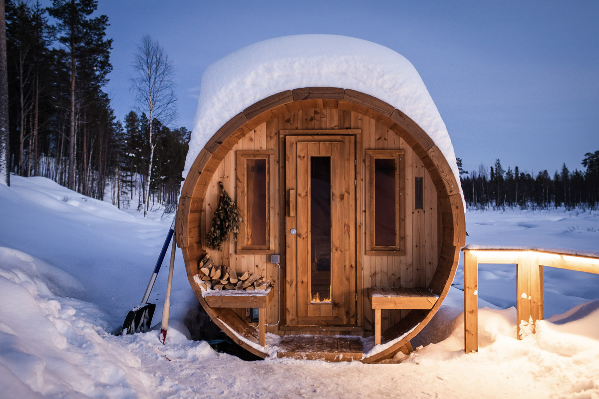 Wooden barrel sauna covered in snow at dusk, with firewood and a birch whisk by the door, Aurora Village, Ivalo