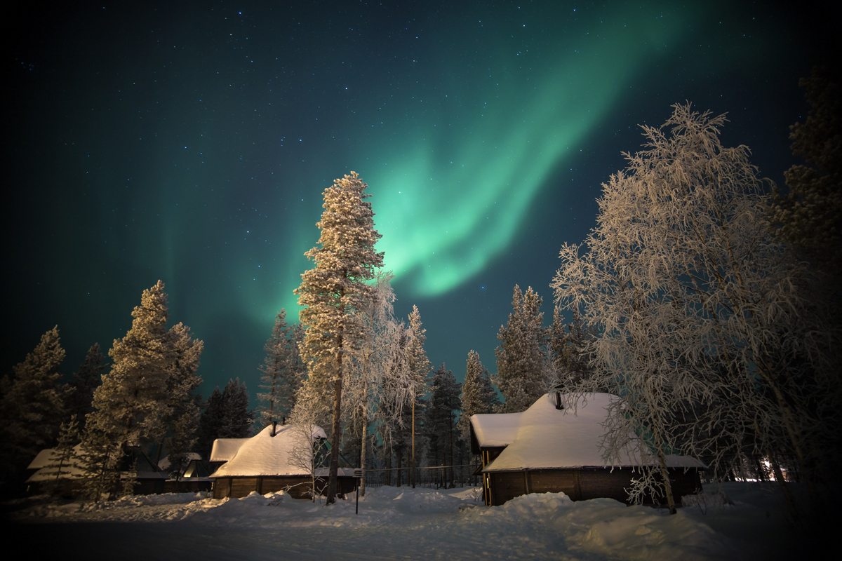 Snow-covered cabins and frost-covered trees under a vivid green northern lights display, Aurora Village, Ivalo