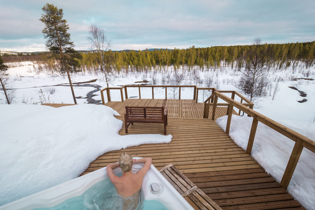 Guest relaxing in an outdoor jacuzzi on a wooden deck with a panoramic view over a snowy landscape, Aurora Village, Ivalo