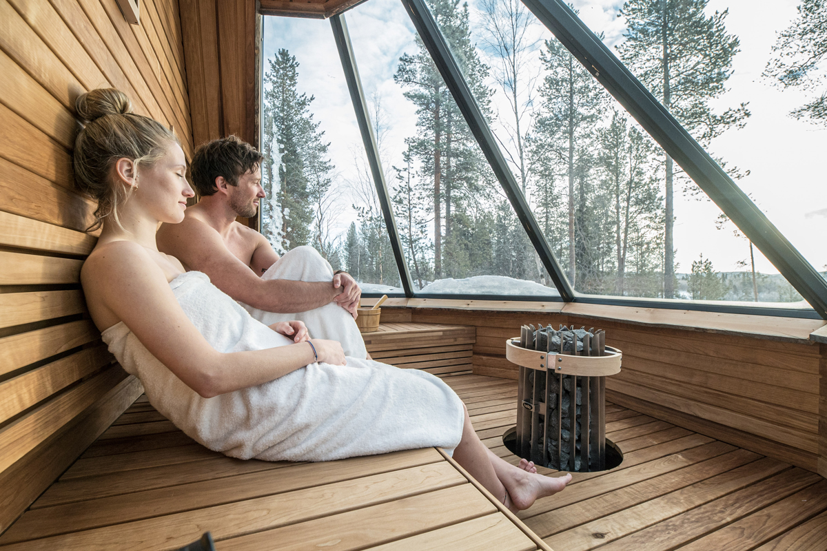 Two guests relaxing in a Finnish sauna with a panoramic glass wall overlooking a snowy forest, Aurora Village, Ivalo