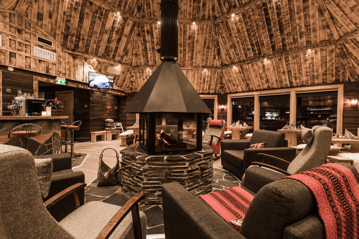 Kota-style restaurant lounge with a central stone fireplace, armchairs and wooden ceiling, Aurora Village, Ivalo