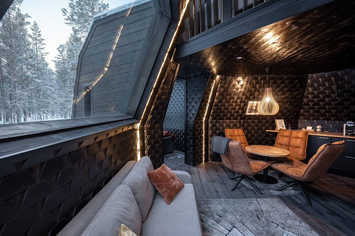 Tower suite interior with angled glass wall, warm lighting, sofa and dining area overlooking a snowy forest, Aurora Village, Ivalo