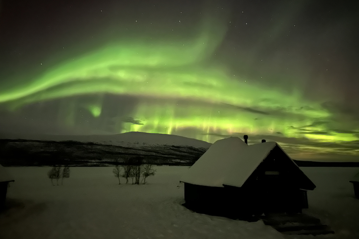 Vivid green northern lights fill the sky above a snow-covered cabin at Arctic Land Adventure, Kilpisjärvi.