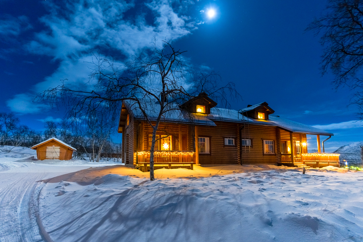 The log cabin lodge glows warmly under moonlight at Arctic Land Adventure, Kilpisjärvi, in winter.