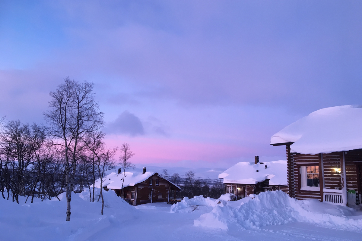 Snow-covered log cabins stand under a pink dusk sky at Arctic Land Adventure, Kilpisjärvi.