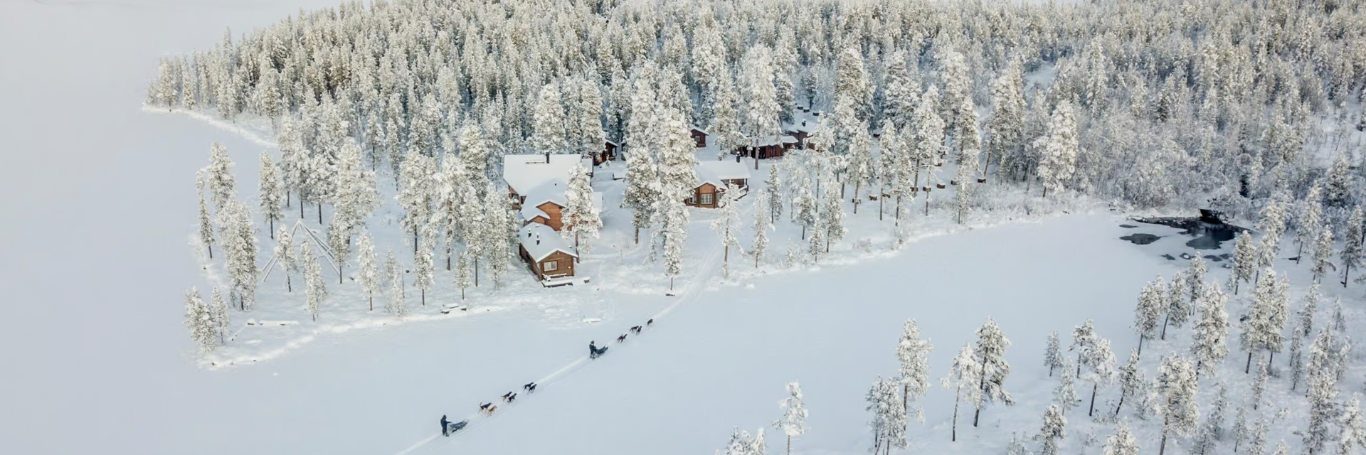Aerial view of a husky sled team approaching Fjellborg Arctic Lodge cabins on a frozen lake in Kiruna.