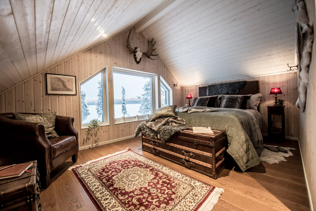 An attic bedroom at Fjellborg Arctic Lodge with a leather armchair, antique trunk, and lake-view windows in Kiruna.