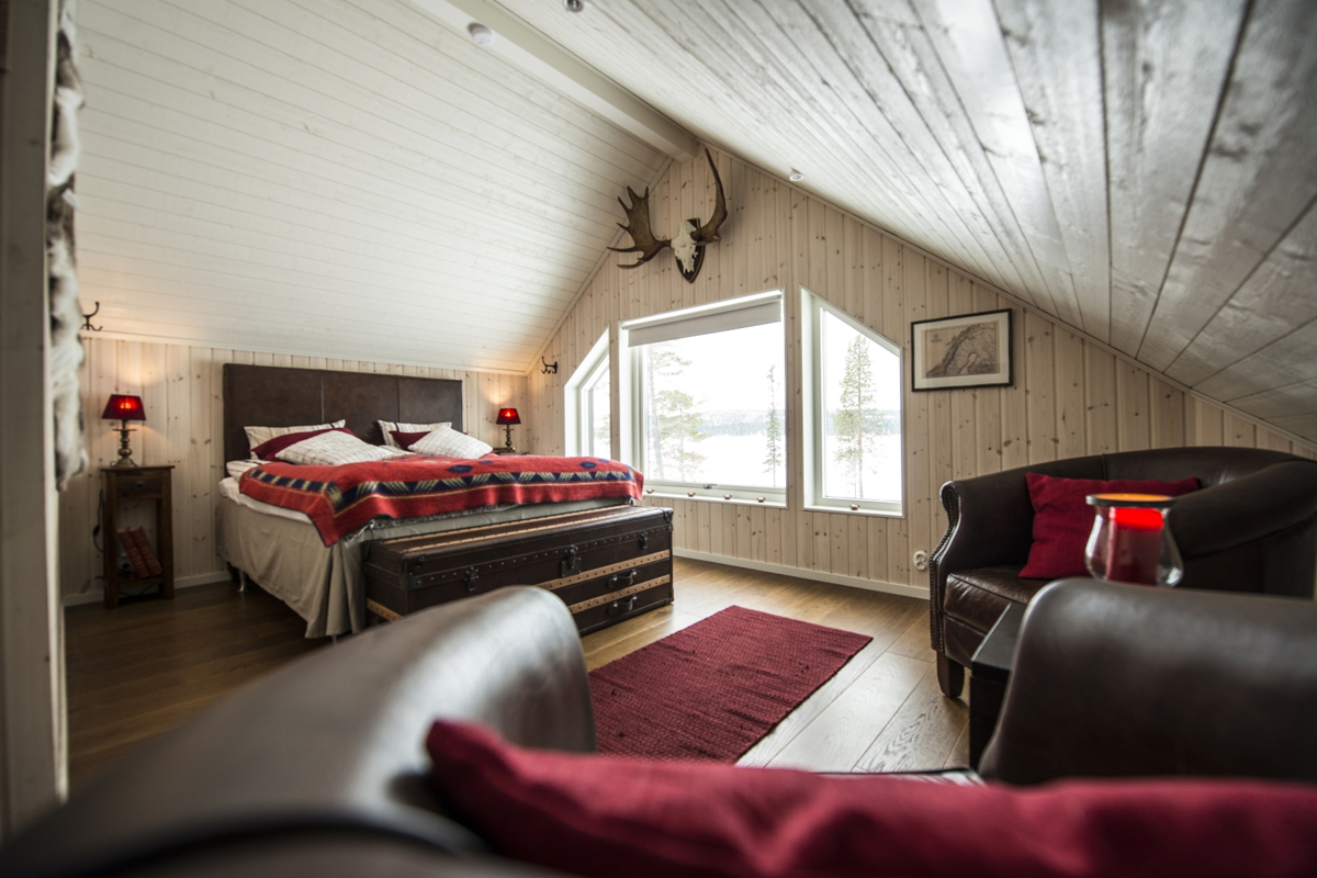 A master bedroom under a sloped wood ceiling with lake views and leather armchairs at Fjellborg Arctic Lodge, Kiruna.