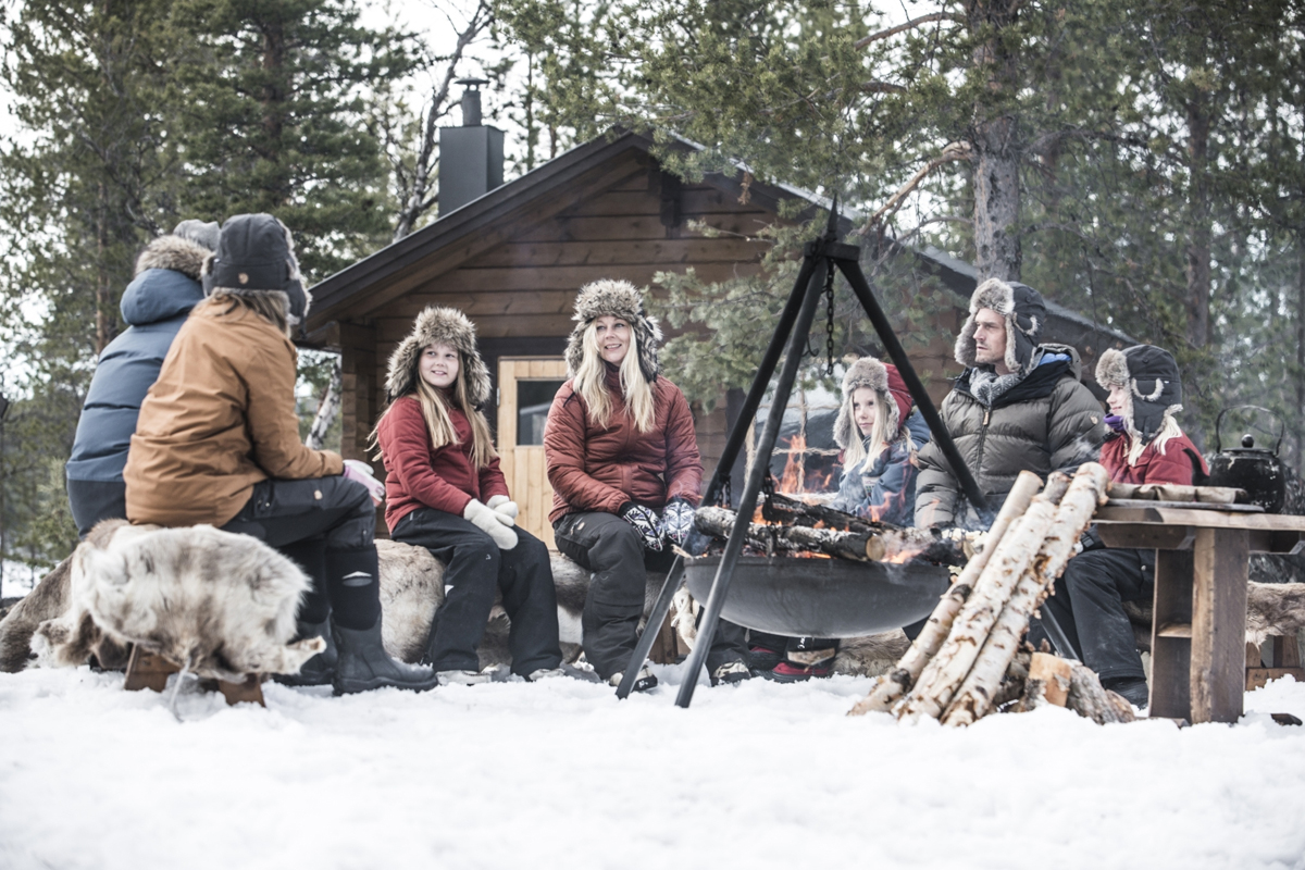 A family gathers around an open fire pit outside a log cabin at Fjellborg Arctic Lodge, Kiruna.
