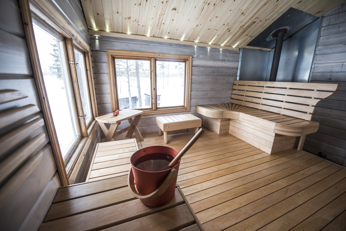 A wooden sauna interior with corner benches and a water bucket overlooking snow at Fjellborg Arctic Lodge, Kiruna.