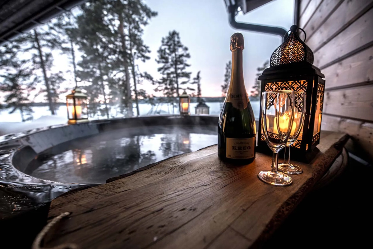 Champagne and glasses beside a steaming outdoor hot tub with lantern light at Fjellborg Arctic Lodge, Kiruna.