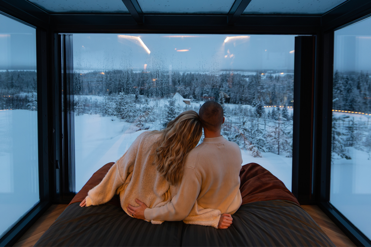 Couple looking out over the forest from the bed inside an Aurora Room at Lumo Resort in Ruka.