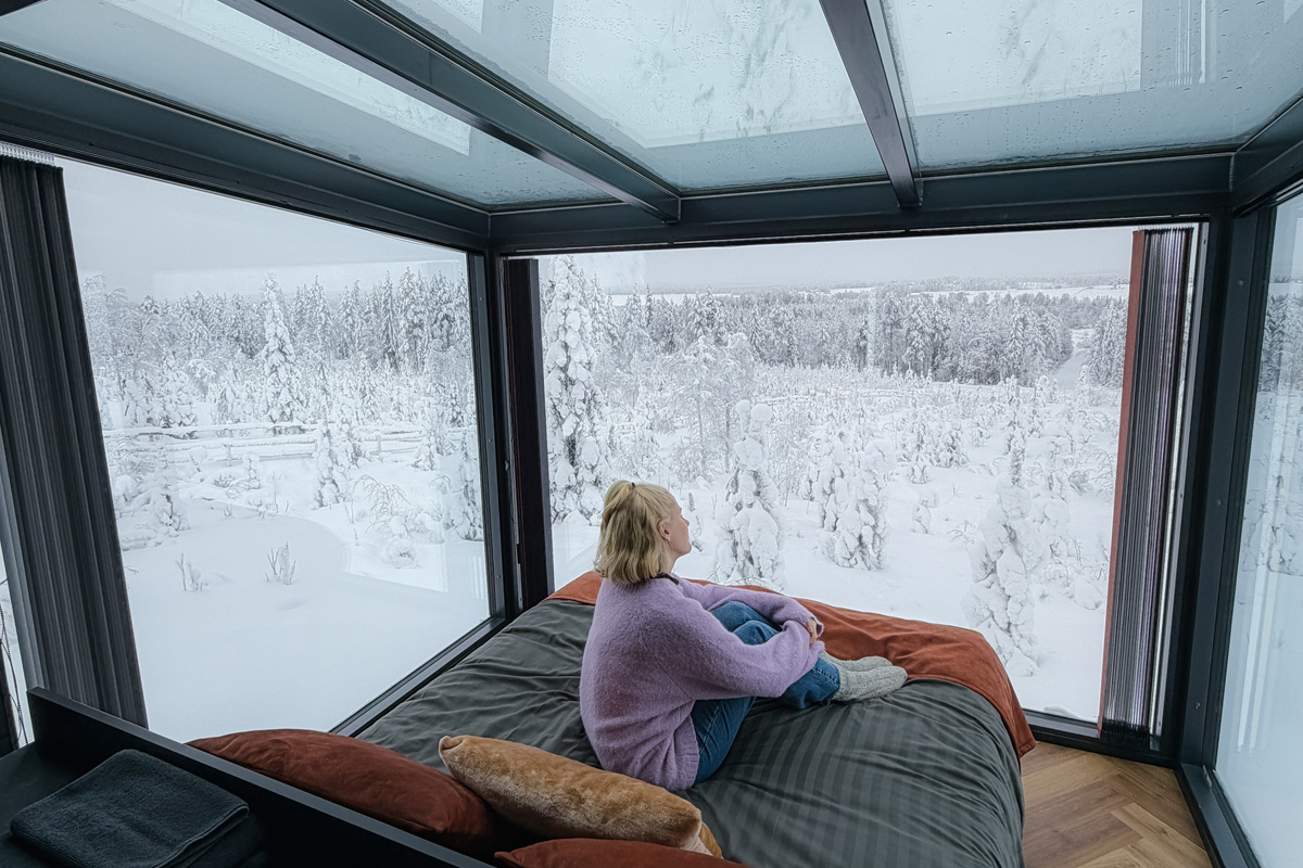 Guest relaxing on the bed inside an Aurora Room at Lumo Resort in Ruka, facing snowy forest.