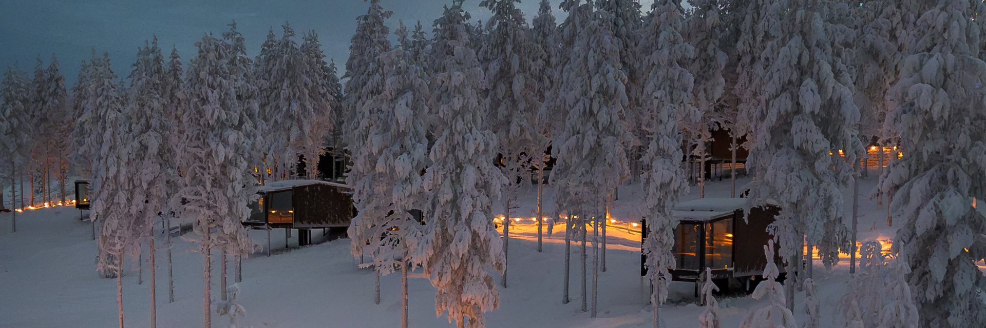 Aurora Rooms among snow covered pine trees at Lumo Resort in Ruka, at dusk.