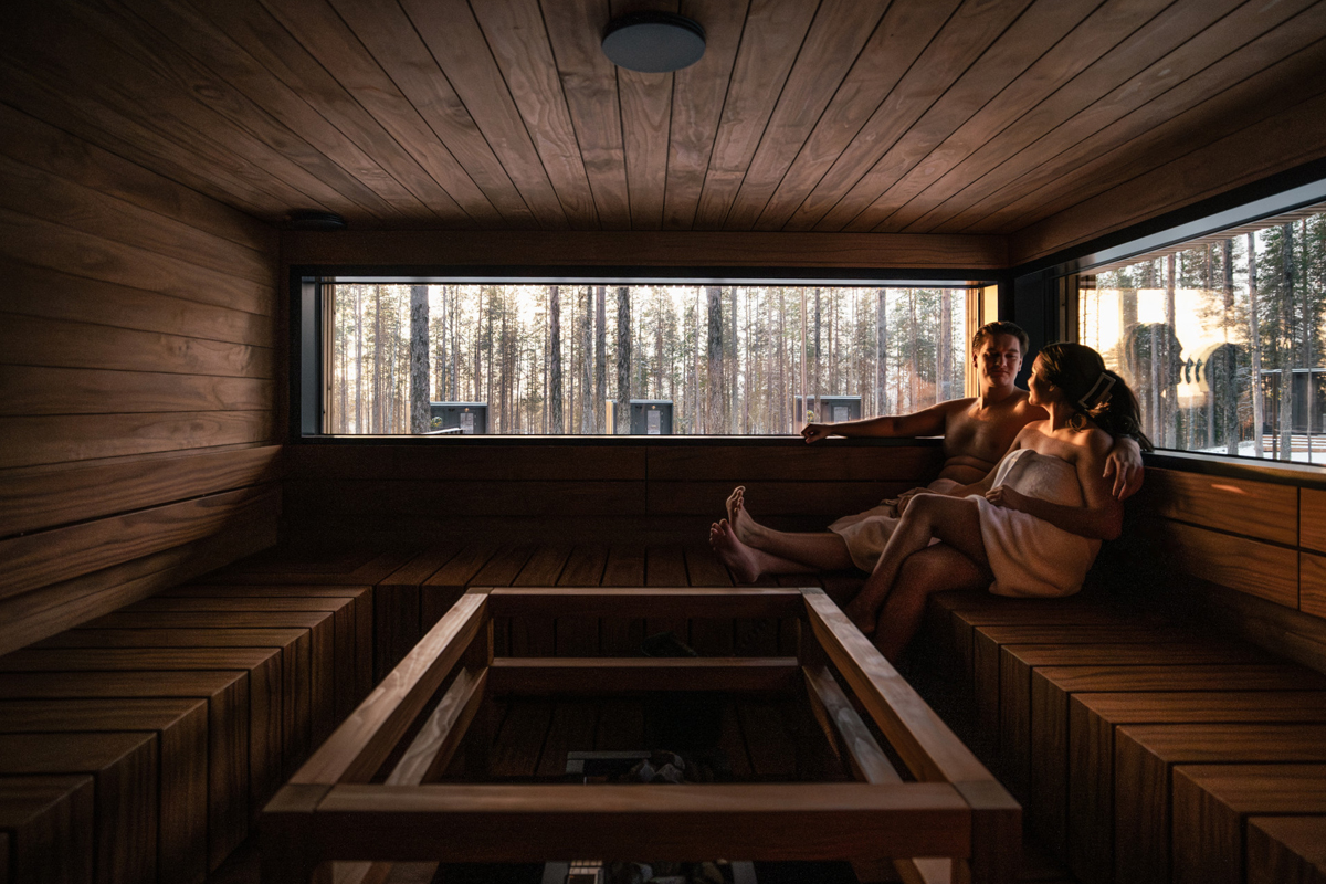 Couple relaxing in a Finnish sauna with a long horizontal window overlooking a pine forest, Olo Resort, Levi