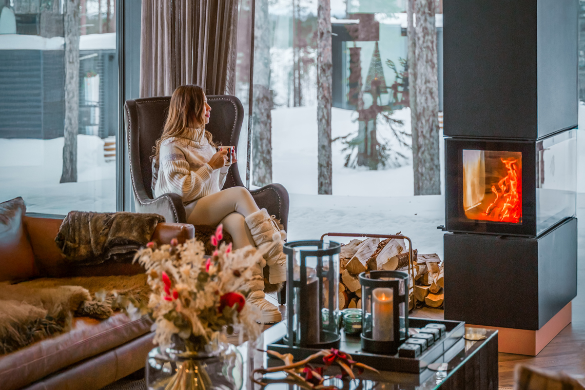 Woman in a cream sweater sitting by a lit fireplace with a snowy forest view through floor-to-ceiling windows, Olo Resort, Levi