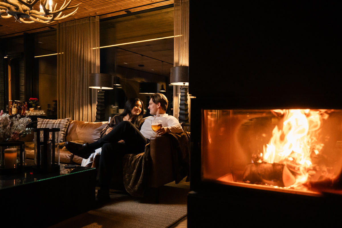 Couple on a leather sofa with drinks beside a glowing fireplace in the main lounge at night, Olo Resort, Levi