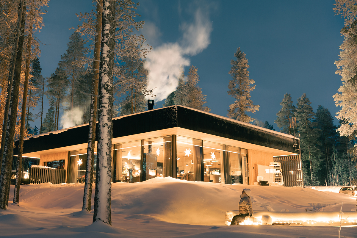 Olo Resort main building lit at night with star decorations visible through floor-to-ceiling windows and smoke rising from the chimney, Levi