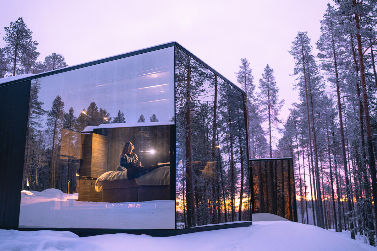 Woman sitting on a bed inside a glass cabin looking up at the sky at sunset, Olo Resort, Levi
