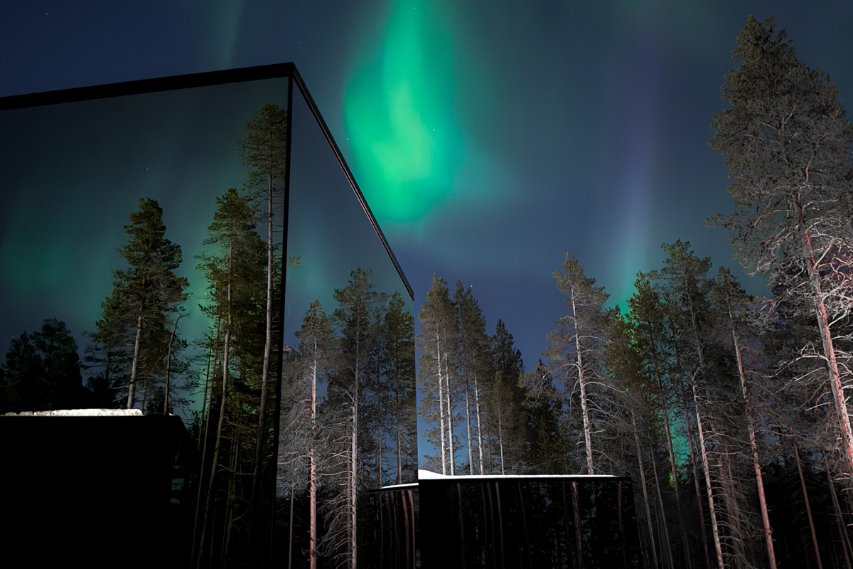 Glass cabin exterior with mirror-like walls reflecting pine trees under a vivid green northern lights display, Olo Resort, Levi
