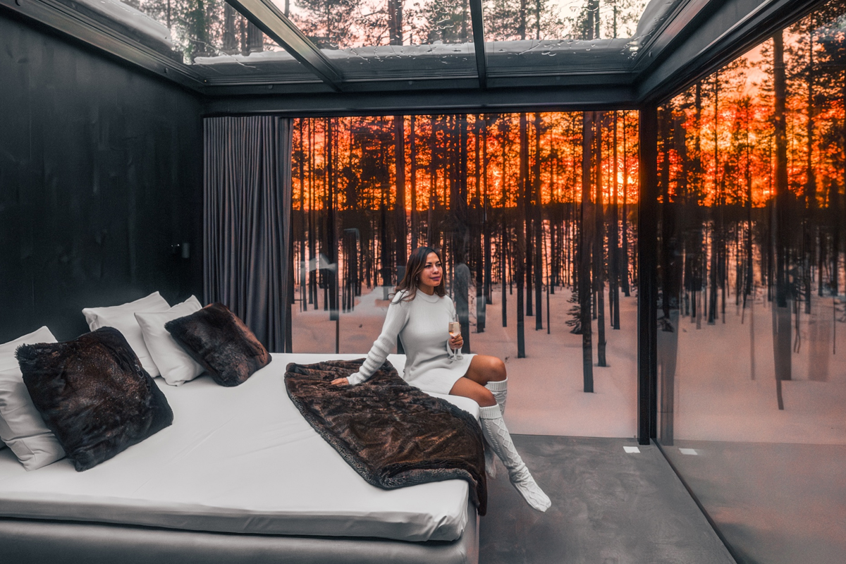 Woman sitting on a bed with a glass of champagne inside a glass cabin at sunset, Olo Resort, Levi