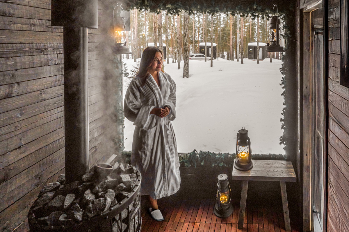 Woman in a bathrobe standing at the open door of an outdoor sauna with steam rising and lanterns, Olo Resort, Levi