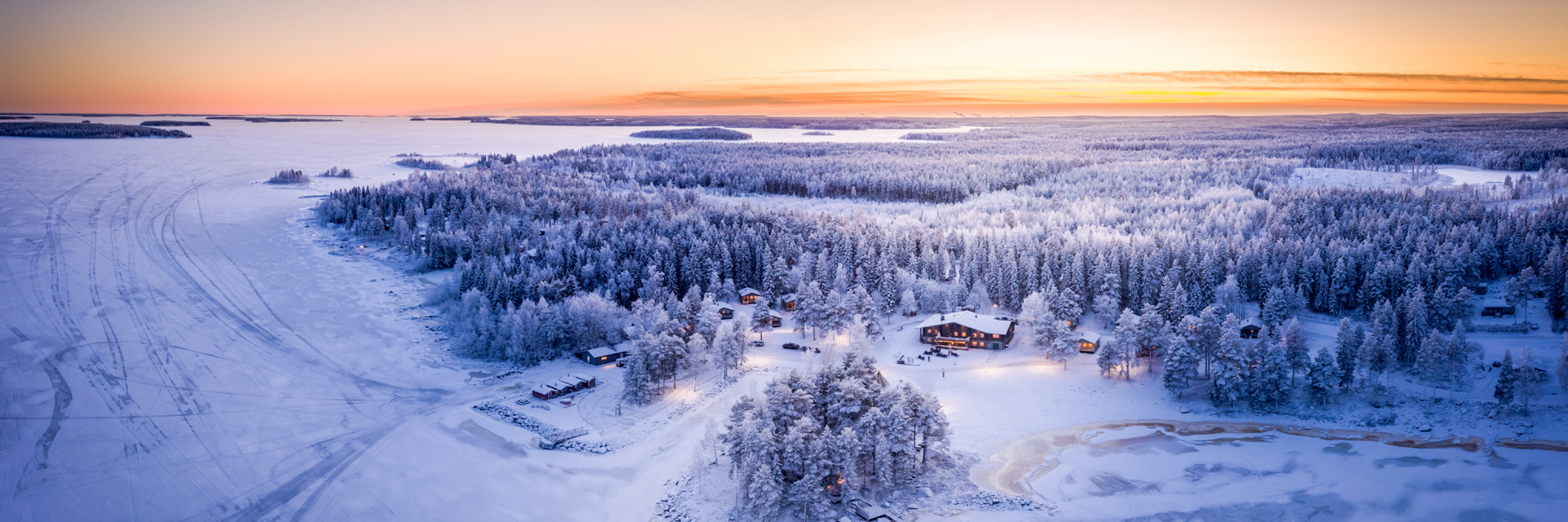 Aerial panoramic view of Brändön Lodge at sunset in winter, with the log cabin restaurant, cabins, and the frozen Luleå archipelago stretching to the horizon.