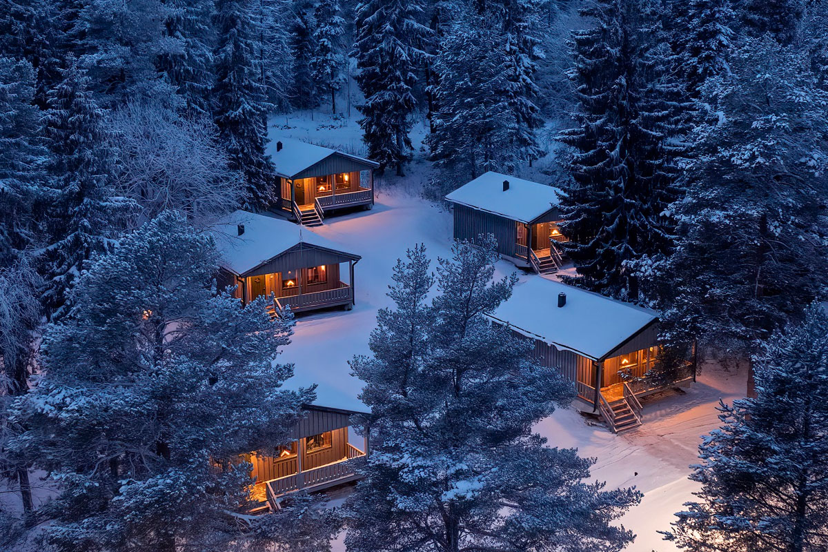 Aerial view of four cabins at Brändön Lodge lit up at night in winter, set among snow-covered trees on the Luleå archipelago coast.