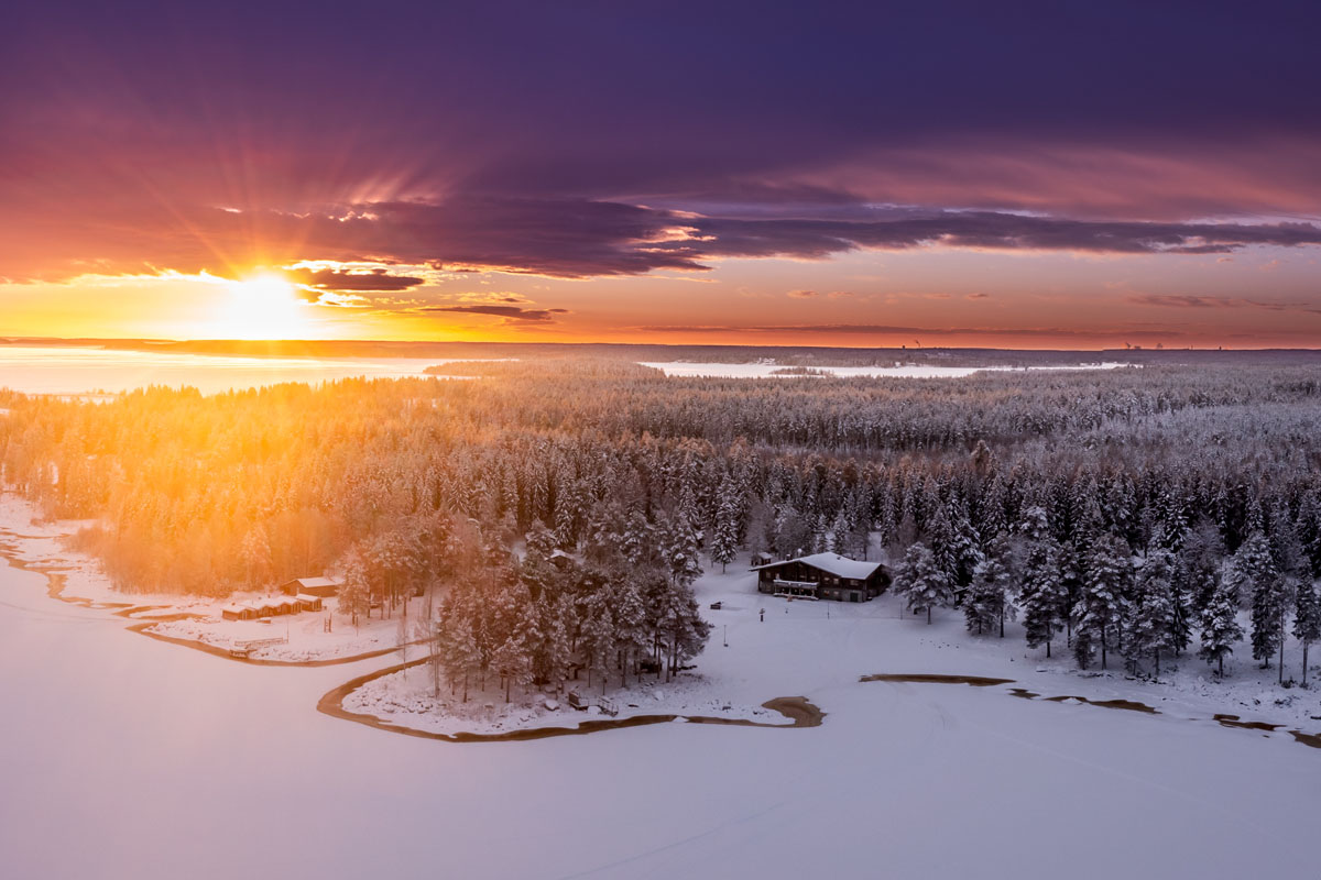 Aerial view of Brändön Lodge at sunset in winter, with the log cabin restaurant visible beside the frozen sea of the Luleå archipelago.