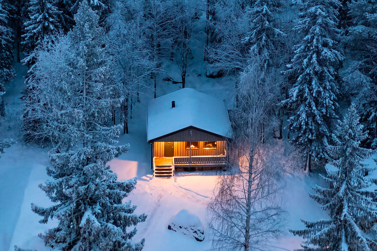 Aerial view of a single cabin at Brändön Lodge at night, glowing warmly among snow-covered trees on the Luleå archipelago coast.