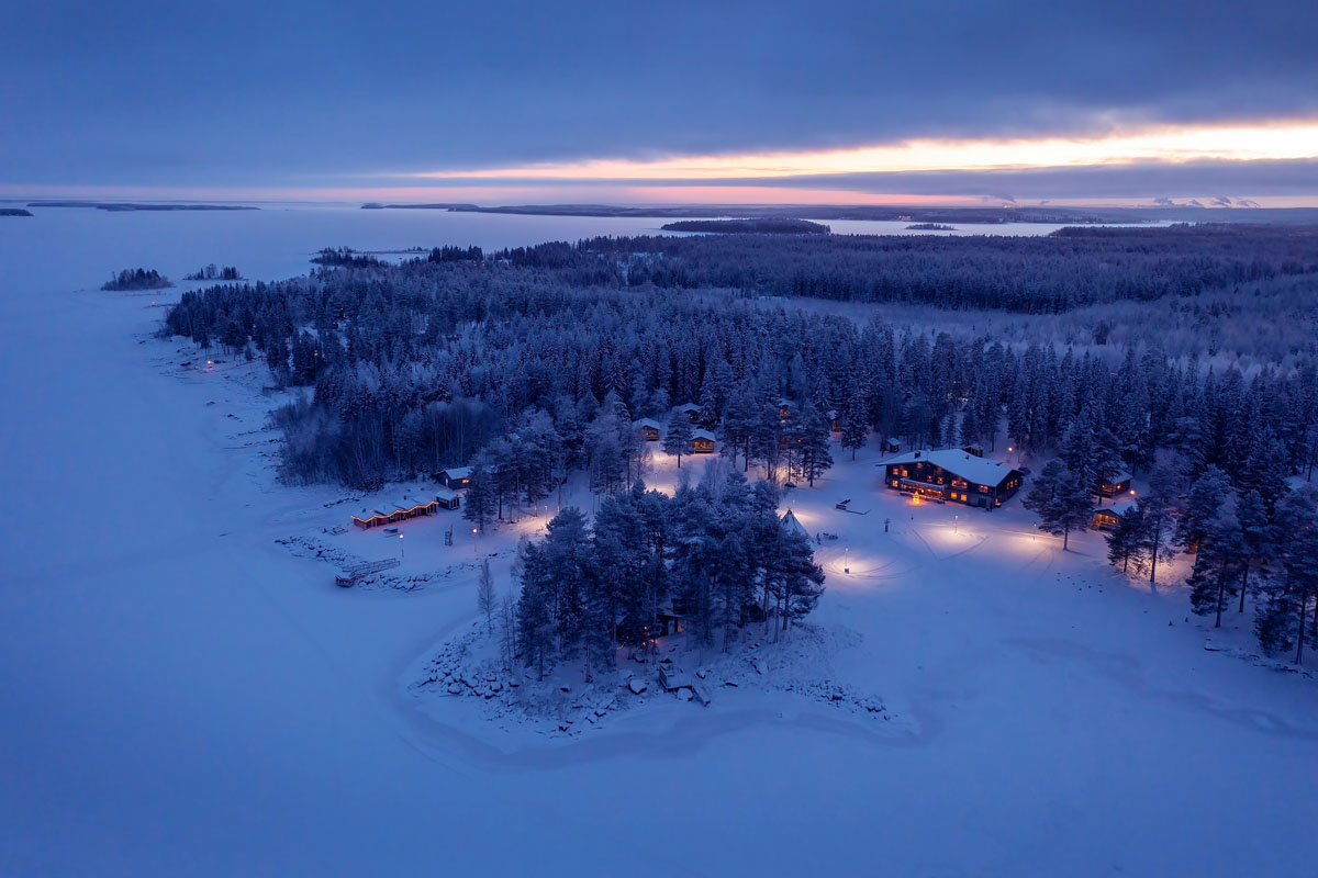 Aerial view of Brändön Lodge at dusk in winter, with cabins lit up among snow-covered trees on the shore of the frozen Luleå archipelago.