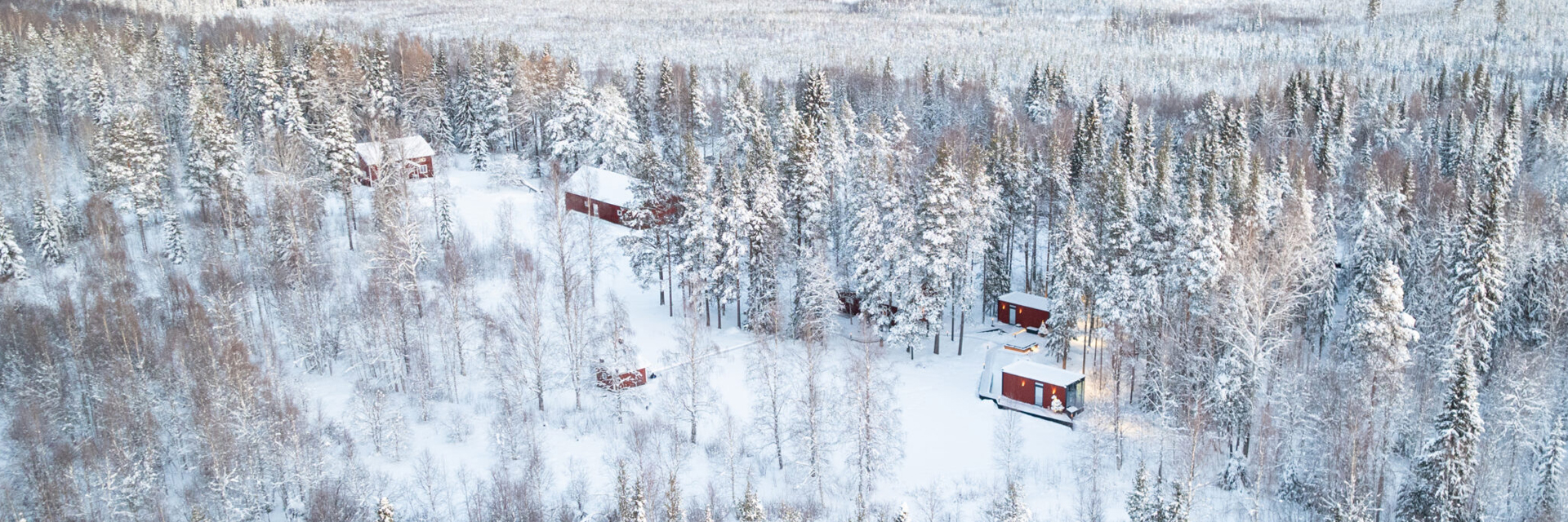 Aerial view of red cabins spread through a snowy pine forest at Loggers Lodge, Harads, Sweden.