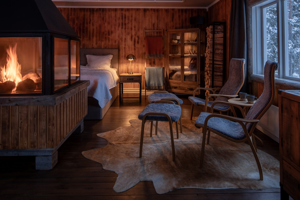 A wood cabin bedroom with fireplace, reindeer hide rug, and armchairs at Loggers Lodge, Harads.