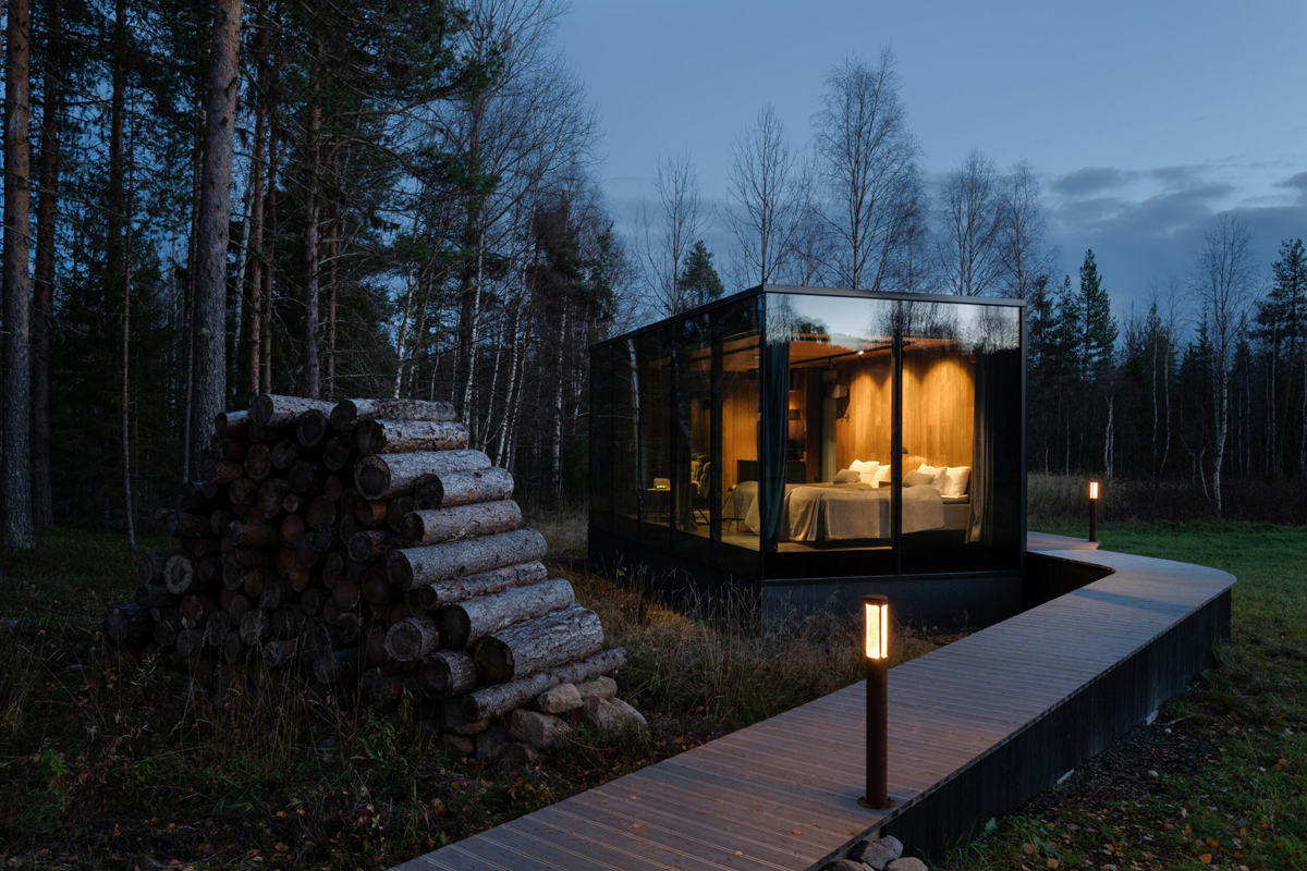A glass pod suite glows at dusk beside stacked logs on a wooden walkway at Loggers Lodge, Harads.