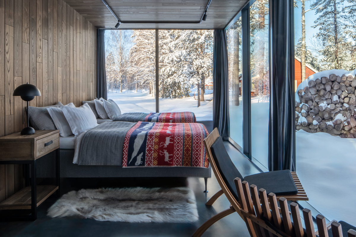 A twin bedroom inside a glass pod at Loggers Lodge overlooks a snowy garden through floor-to-ceiling windows.