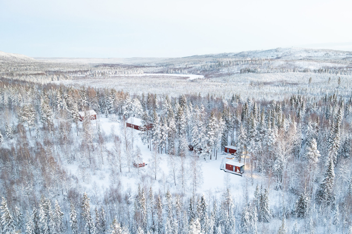 Aerial view of several red cabins scattered across a snowy forest clearing at Loggers Lodge, Harads.