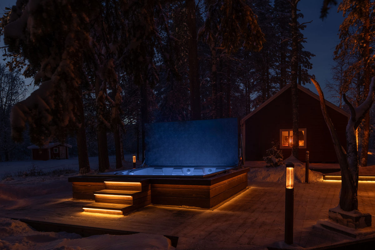 An outdoor hot tub glows at dusk on a wooden deck beside the main lodge at Loggers Lodge, Harads.