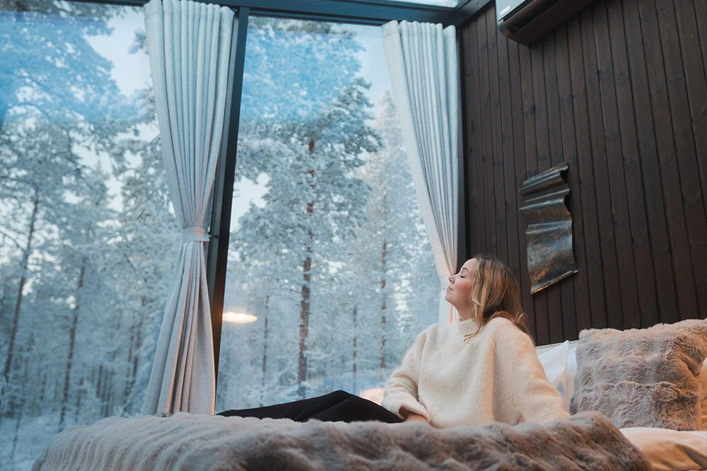 Woman sitting on a fur-covered bed in a glass igloo with a panoramic view of a snowy forest, Halo Igloos, Rovaniemi