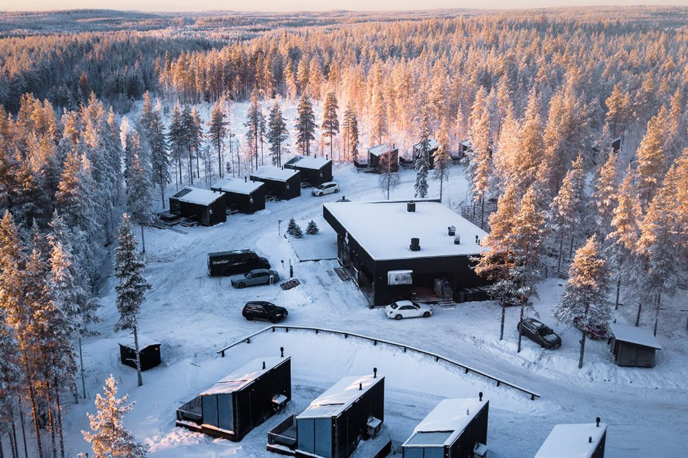 Aerial view of Halo Igloos with dark cabins and main building set among frost-covered trees at sunrise, Rovaniemi