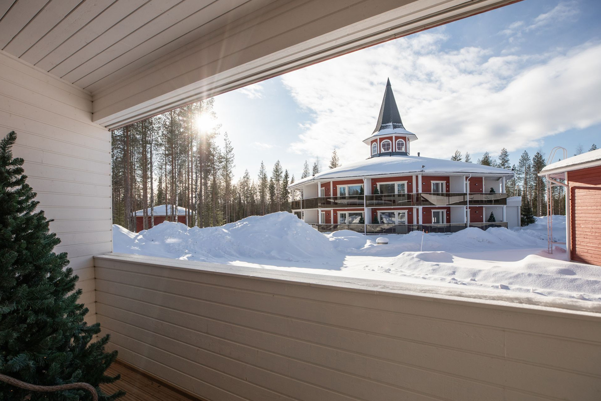 Balcony view of a snow-covered main building with a pointed tower at Santa Claus Holiday Village on a sunny winter day, Rovaniemi