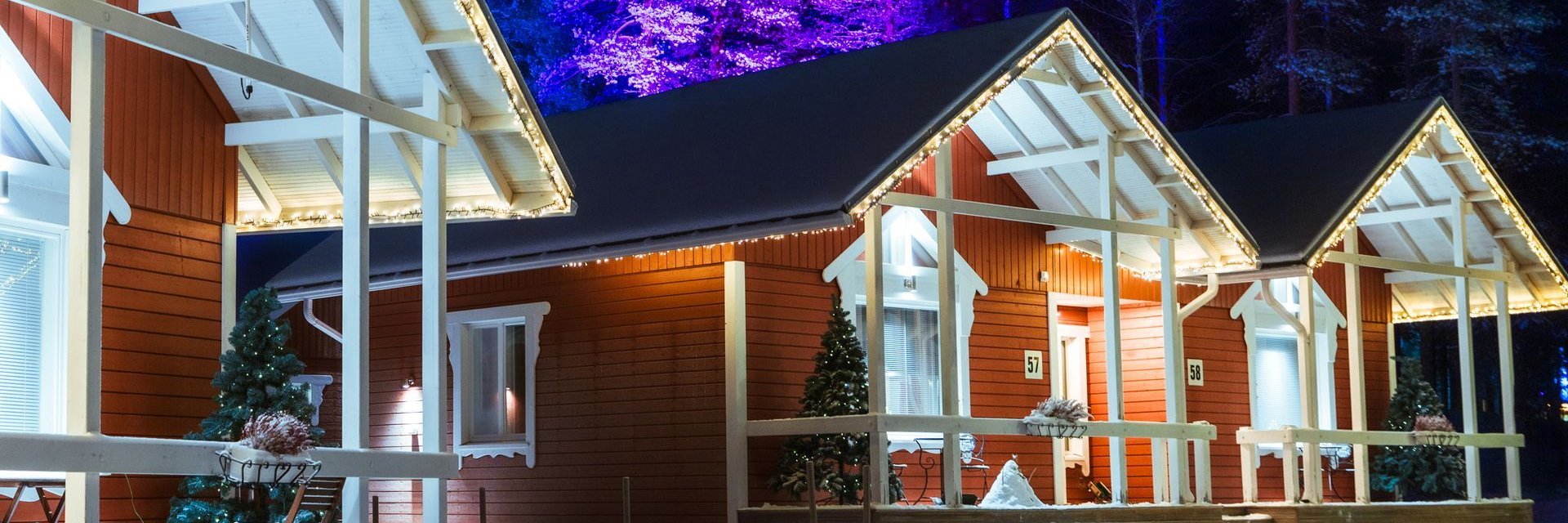 Red classic cottages lit with Christmas lights at night, with trees illuminated in purple and blue, Santa Claus Holiday Village, Rovaniemi