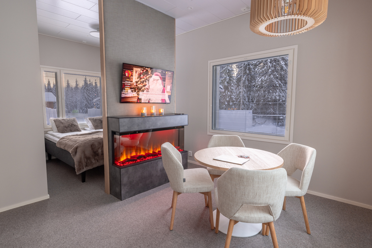 Dining area with fireplace and TV in a glass igloo cottage, with a sleeping area visible beyond, Santa Claus Holiday Village, Rovaniemi
